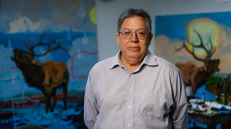 Norman Akers standing in front of two of his paintings that are displayed on the wall behind him