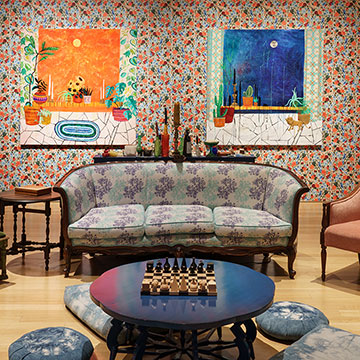 A couch and four assorted accent chairs arranged around a coffee table with a chess set on top. The couch and chairs are placed in a gallery in front of a wall that has patterned wallpaper and two large paintings by Amy Kligman.