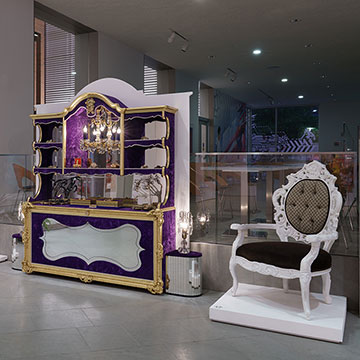 A view of an art installation at the Nerman Museum by Carlos Rolon showcasing a purple shelf, a chair and a large wall hanging that says ALL WE HAVE IS RIGHT NOW
